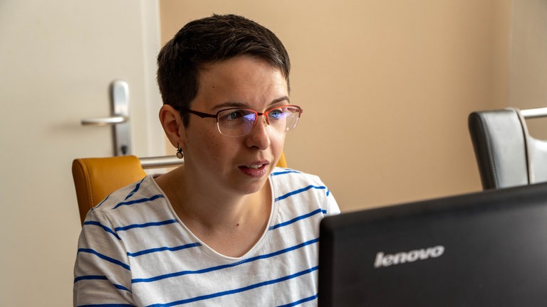 Eine Frau im gestreiften T-Shirt mit kurzen schwarzen Haaren und Brille sitzt vor einem Laptop und schaut auf diesen.