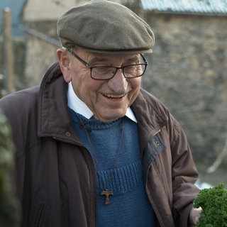 Ein alter Mann mit Mütze und Brille lacht. Albert ist seit 65 Jahren Priester im Hohen Westerwald.