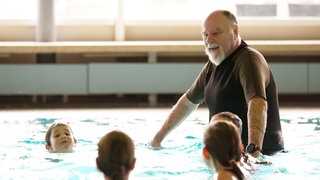 Ein Mann mit weißem Bart und Glatze ist mit drei Kindern im Schwimmbecken