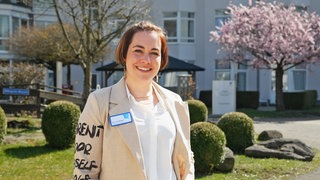 Frau mit kurzen braunen Haaren und Blazer steht vor Bäumen und Büschen.