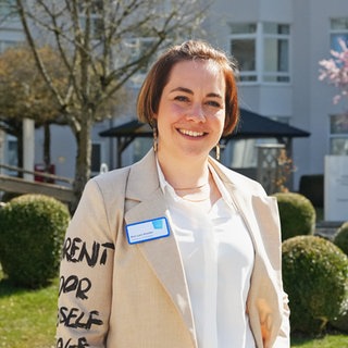 Frau mit kurzen braunen Haaren und Blazer steht vor Bäumen und Büschen.