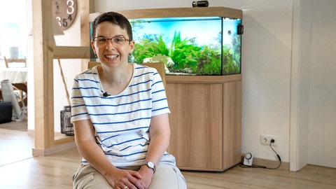 Eine Frau im gestreiften T-Shirt mit kurzen schwarzen Haaren und Brille sitzt vor einem Aquarium und lächelt.