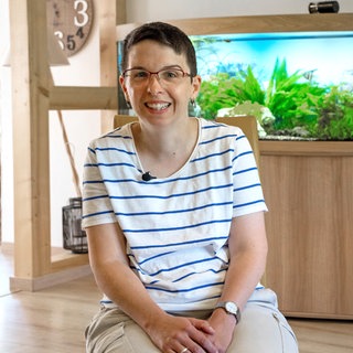 Eine Frau im gestreiften T-Shirt mit kurzen schwarzen Haaren und Brille sitzt vor einem Aquarium und lächelt.