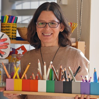 Frau mit Brille, baunen Pullover und Halskette trägt ein Brett mit Holzfarbstiften in der Hand.