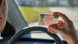 Ein junger Mann sitzt im Auto und hält seinen Führerschein vor das Lenkrad