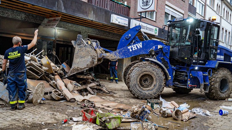 Schutt und Müll, der in Folge einer Überschwemmung auf einer Straße liegt, wird von einem THW-Bagger beseitigt