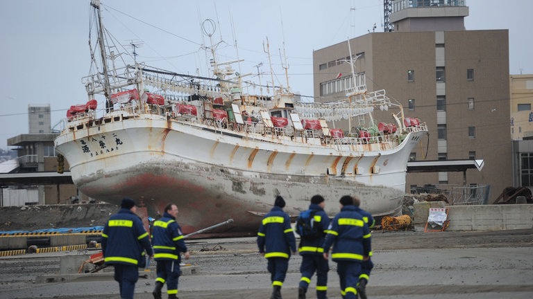 Im Vordergrund laufen sechs Helfer des THW in blauer Einsatzkleidung. Im Hintergrund steht ein großes Schiff, das durch den Tsunami an Land gespült wurde.