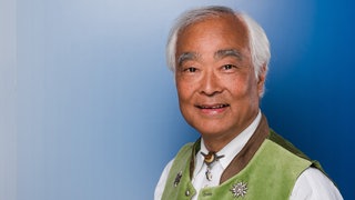 Der Japanische Jodel-Künstler Take Ischi in einer bayerischen Tracht vor einem blauen Hintergrund