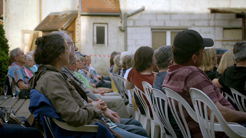 Theaterbesucher sitzen auf Plastikstühlen im Hof