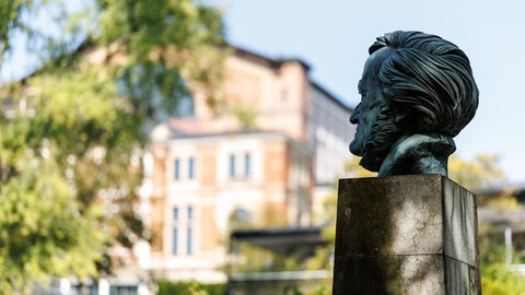Wagner-Büste im Profil, im Hintergund verwaschen das Bayreuther Festspielhaus.