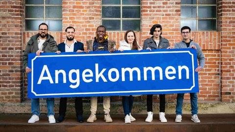 Sechs geflüchtete Menschen halten ein Schild „Angekommen“ in der Hand