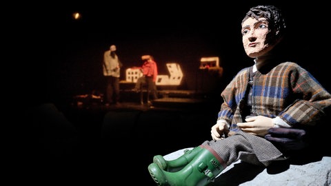 Szene aus dem Puppentheater: große Frauenpuppe sitzt vorne rechts