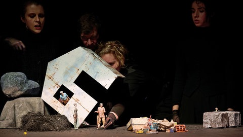 Vier Puppenspieler spielen mit kleinen Figuren eine Szene, die in einem Haus spielt.