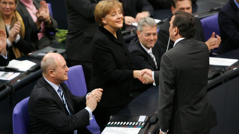 Angela Merkel im Bundestag, Gerhard Schröder schüttelt ihr die Hand, Gregor Gysi applaudiert daneben 