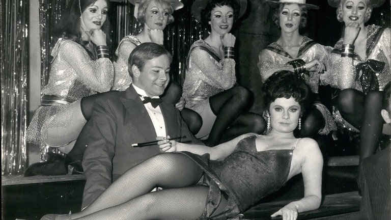 Judi Dench räkelt sich im Abendkleid mit Netztrümpfen und Zigarettenspitze vor David Bexon (Conférencier) und Ensemble-Mitgliedern