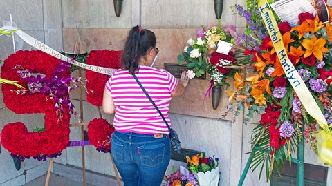 Noch heute pilgern Fans aus der ganzen Welt am 1. Juni — ihrem Geburtstag — zum Grab von Marilyn Monroe. 2021 wäre die Schauspielikone 95 Jahre alt geworden: Eine junge Frau im gestreiften T-Shirt und in Jeans steht auf einem Friedhof vor einer Sargnische aus grau-braunem Marmor. Sie berührt eine Platte auf der ein Metallschild mit dem Namen Marilyn Monroe eingelassen ist. An der Wand stehen Blumenkränze, eine 95 aus Rosen und weitere Trauerbekundungen. Ein Flor mit der Aufschrift 'Immortal Marilyn' hängt quer über einem Kranz.