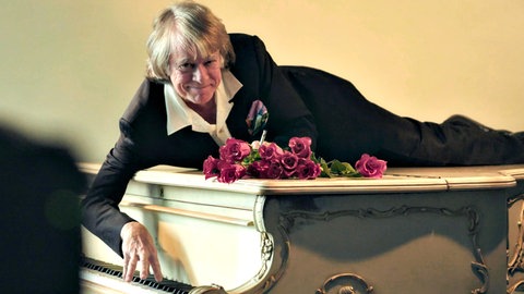 Helge Schneider liegt auf einem weißen Flügel, vor ihm ein Strauß roter Rosen