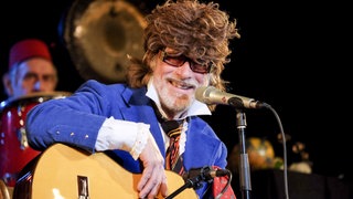 Helge Schneider mit großer Perücke und Sonnenbrille bei einem Konzert mit Gitarre