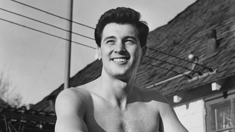 Rock Hudson lehnt oberkörperfrei an einem Zaun. Bild von 1951