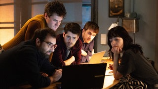 "The Kollective - Der Absturz": Etienne (Grégory Montel), Joshua (Gregg Sulkin) und Aaron (Felix Mayr) sitzen in einem Zimmer an einem kleinen Tisch. Dahinter steht Lucas (Gijs Blom), während Delia (Céline Buckens) ihnen gegenübersitzt. Alle fünf betrachten etwas auf einem Laptop.