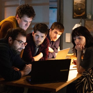 "The Kollective - Der Absturz": Etienne (Grégory Montel), Joshua (Gregg Sulkin) und Aaron (Felix Mayr) sitzen in einem Zimmer an einem kleinen Tisch. Dahinter steht Lucas (Gijs Blom), während Delia (Céline Buckens) ihnen gegenübersitzt. Alle fünf betrachten etwas auf einem Laptop.