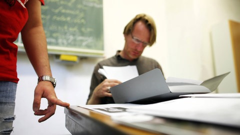 Ein Lehrer sitzt vor einem Schüler und blickt auf dessen Arbeit. Lehrkräfte versuchen Kinder mit Migrationshintergrund leicht besser zu bewerten.