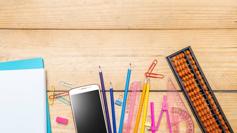 Symbolbild eines Handys inmitten von Schulutensilien auf einem Tisch, tags: Smartphone-Verbot Schule Smartphones