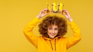 Eine junge Frau mit Locken setzt sich lächelnd eine Krone auf den Kopf