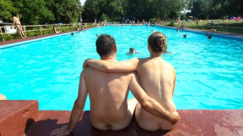 Ein nacktes Paar von hinten sitzt im Freibad am Rand eines Schwimmbeckens.