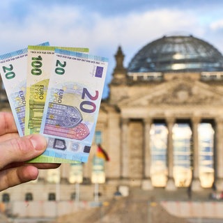 Eine Hand hält Euro-Geldscheine vor dem Reichstagsgebäude in Berlin.