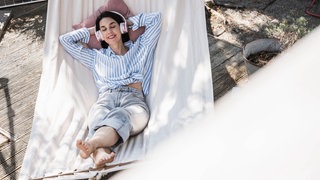 Eine lächelnde Frau liegt mit Kopfhörern und geschlossenen Augen in einer Hängematte.