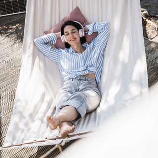 Eine lächelnde Frau liegt mit Kopfhörern und geschlossenen Augen in einer Hängematte.