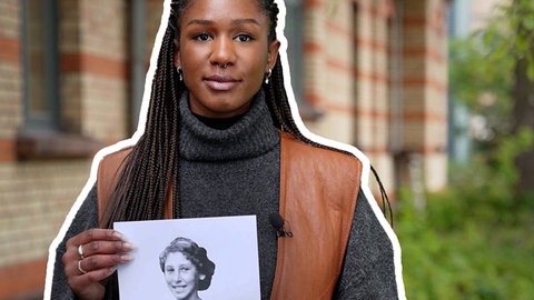 Eine Frau hält ein Schwarz-Weiß-Foto in die Kamera mit einem lächelnden Mädchen darauf, im HIntergrund ist ein Backsteingebäude zu sehen