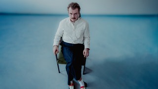 Artur Weigandt sitzt auf einem Stuhl mitten in einem Raum