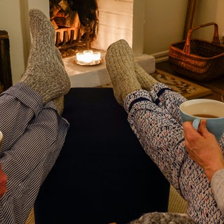 A couple sit together drinking tea with wool socks in front of fireplace. Symbolfoto.
