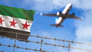 Symbolbild zu Abschiebungen nach Syrien: Ein Stacheldrahtzaun, eine syrische Flagge und ein Flugzeug 