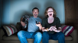 Ein Paar mittleren Alters und ihr Hund lachen über den Fernseher. Symbolfoto.