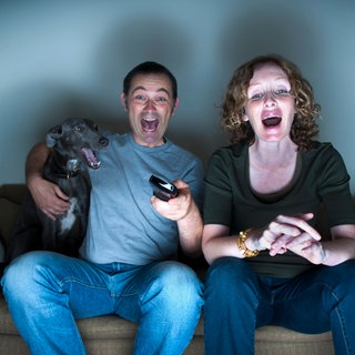 Ein Paar mittleren Alters und ihr Hund lachen über den Fernseher. Symbolfoto.