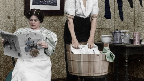 Eine Frau liest die Zeitung während ein Mann die Wäsche wäscht