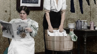 Eine Frau liest die Zeitung während ein Mann die Wäsche wäscht