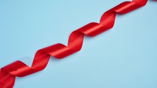Ein rotes Satinband, zu einer Spirale gedreht, ist diagonal auf einem blauen Hintergrund platziert. Symbolfoto.