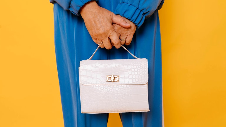 Frau in blauem Outfit mit weißer Handtasche vor gelbem Hintergrund. Symbolfoto.