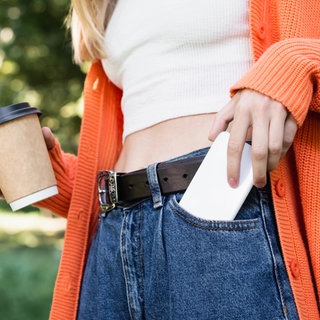 Ausschnitt einer Frau, die einen Pappbecher hält und gleichzeitig ihr Smartphone aus der Tasche holt. Symbolfoto.