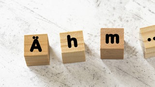 Vier Holzwürfel bilden das Füllwort "Ähm" auf einer weißen Zementplatte. Symbolfoto.
