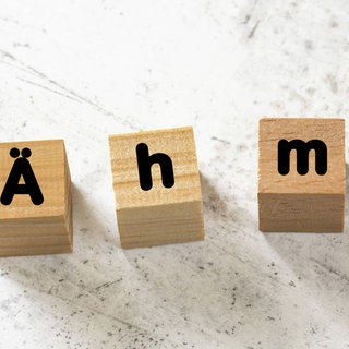 Vier Holzwürfel bilden das Füllwort "Ähm" auf einer weißen Zementplatte. Symbolfoto.