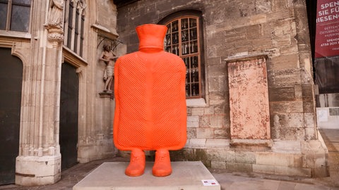 Eine überdimesional große orangefarbene Wärmflasche auf zwei Beinen steht vor dem Stephansdom in Wien.