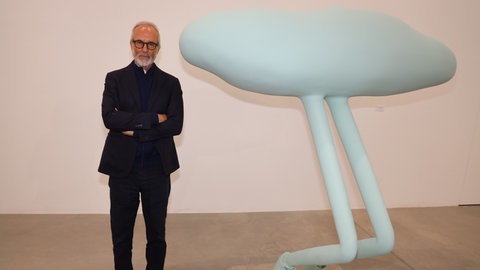 Künstler Erwin Wurm steht neben einer mintfarbenen Skulptur mit überlangen Beinen.
