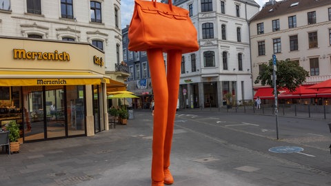 Eine orangefarbene riesige Handtasche auf langen Beinen als Skulpur scheint in einer Einkaufsstraße auszuschreiten. 