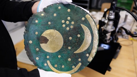Die Himmelsscheibe von Nebra wird in die Kamera gehalten, im Hintergrund ist ein Mikroskop zu sehen. 