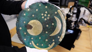 Die Himmelsscheibe von Nebra wird in die Kamera gehalten, im Hintergrund ist ein Mikroskop zu sehen. 